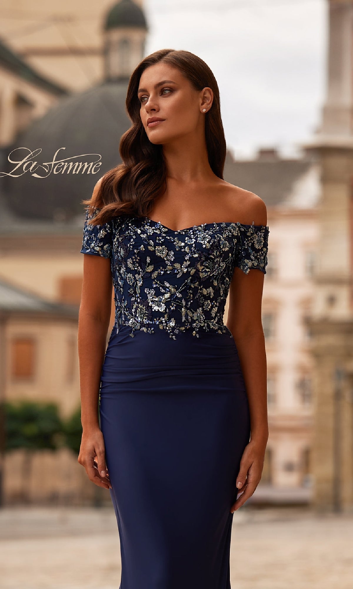Outdoors in the city, a woman wears the Sequin-Bodice Long Navy Prom Dress (La Femme 32739)—an off-the-shoulder gown with a sparkling bodice that offers a perfect blend of sophistication and modern style.